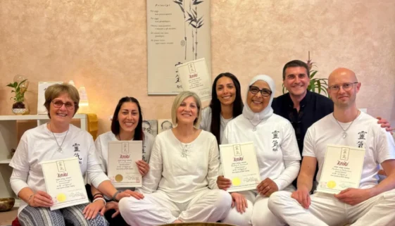 Gruppo di persone sedute in cerimonia di reiki, alcuni tengono certificati di reiki primo livello, ambiente sereno con campane e decorazioni zen.