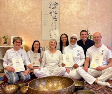 Gruppo di persone sedute a terra durante una sessione di meditazione o formazione Reiki, con certificati in mano e grandi campane tibetane davanti. Insieme indossano abiti bianchi, in un ambiente accogliente e zen. Incorporando il termine corso reiki.
