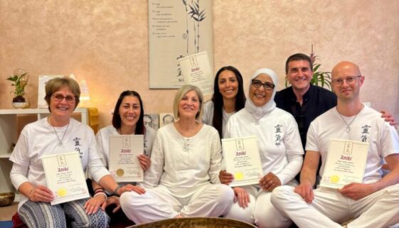 Gruppo di persone sedute a terra durante una sessione di meditazione o formazione Reiki, con certificati in mano e grandi campane tibetane davanti. Insieme indossano abiti bianchi, in un ambiente accogliente e zen. Incorporando il termine corso reiki.