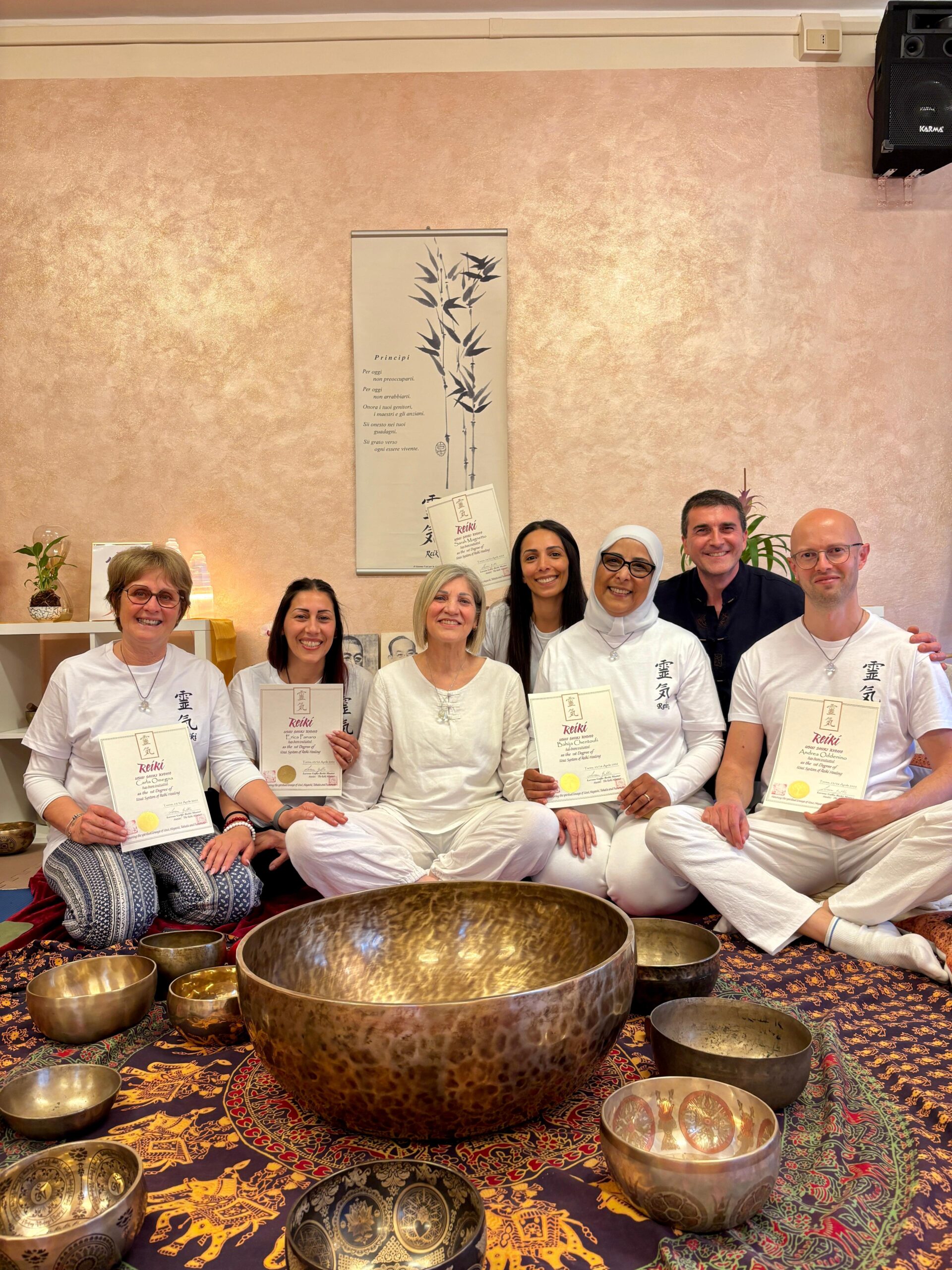 Gruppo di persone sedute a terra durante una sessione di meditazione o formazione Reiki, con certificati in mano e grandi campane tibetane davanti. Insieme indossano abiti bianchi, in un ambiente accogliente e zen. Incorporando il termine corso reiki.