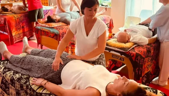 Donna distesa sul lettino, mentre operatore appoggia le mani sul corpo; varie persone ricevendo trattamenti in una sala colorata, ampi lettini e massaggiatori. come funziona il reiki.