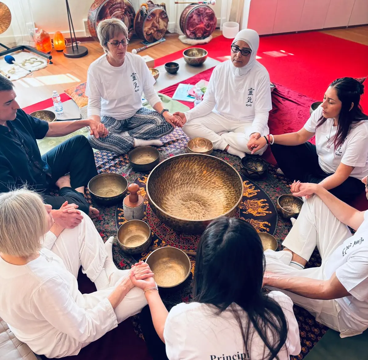 Gruppo di persone sedute in cerchio su tappeto, con strumenti rituali al centro durante una sessione di meditazione Reiki, indossano abiti bianchi.